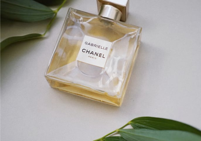 A bottle of Gabrielle Essense perfume laying flat against a table with green leaves above and below it